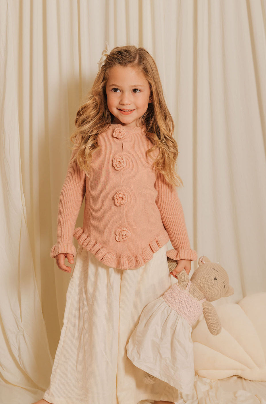 Young girl in a pink cardigan and white skirt holding a teddy bear against a beige curtain background
